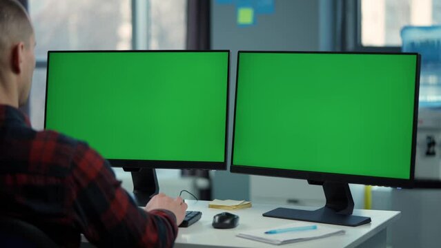 Young Man Working On Computer With Two Green Screen Mock Ups Sitting At Desk In Office