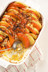 Sliced and layered potato and pumpkin casserole in a white baking form on marble background, topped with roasted onions, feta cheese crumbs and whipped cream