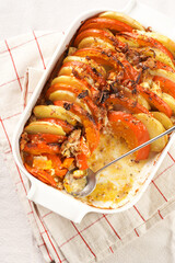 Sliced and layered potato and pumpkin casserole in a white baking form on marble background, topped with roasted onions, feta cheese crumbs and whipped cream