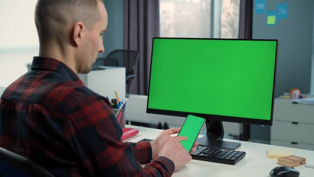 Man Uses Phone With Green Mock Up Screen on Background Green Screen Monitor in Office