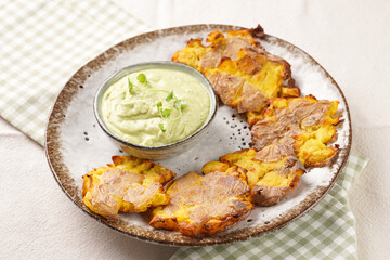 Trendy comfort snack crispy roasted crushed potatoes on a round plate with creamy avocado and garlic dip on a green checkered kitchen towel