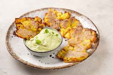 Trendy comfort snack crispy roasted crushed potatoes on a round plate with creamy avocado and garlic dip on a beige colored table cloth