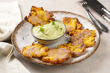 Trendy comfort snack crispy roasted crushed potatoes on a round plate with creamy avocado and garlic dip on a beige colored table cloth
