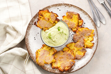 Trendy comfort snack crispy roasted crushed potatoes on a round plate with creamy avocado and garlic dip on a beige colored table cloth, top view