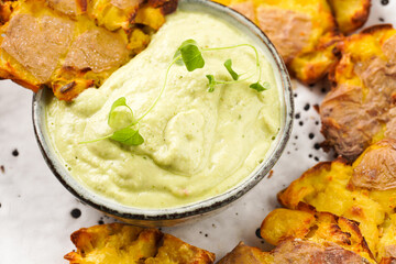 Trendy comfort snack crispy roasted crushed potatoes on a round plate with creamy avocado and garlic dip on a beige colored table cloth, close up