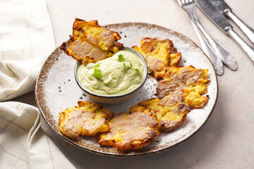 Trendy comfort snack crispy roasted crushed potatoes on a round plate with creamy avocado and garlic dip on a beige colored table cloth