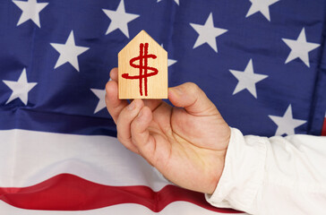 Against the background of the USA flag, a man's hand holds a wooden house with the image of the dollar symbol.
