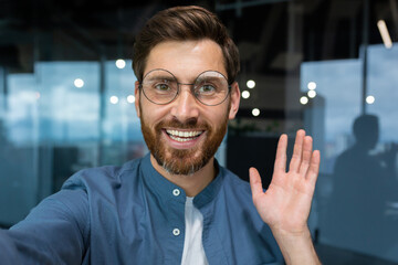 Portrait of successful businessman in casual shirt and glasses man looking at smartphone camera and smiling, camera view video call talking with friends inside office remotely.