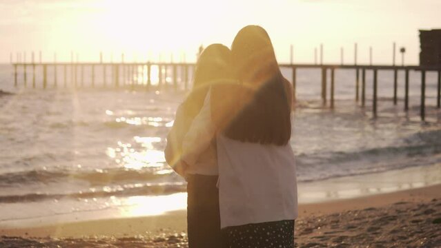 Photographer takes pictures An adult daughter hugs her mother at sunset by the sea. Back view, sun glare