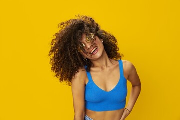 Woman with curly afro hair in a blue T-shirt on a yellow background dancing flying hair with sunglasses yellow, hand signs, look into the camera, smile with teeth and happiness, copy space