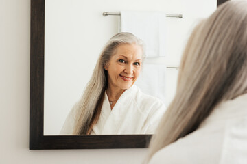Older woman with long beautiful hair admiring herself in a mirror. Senior female in a bathroom in...