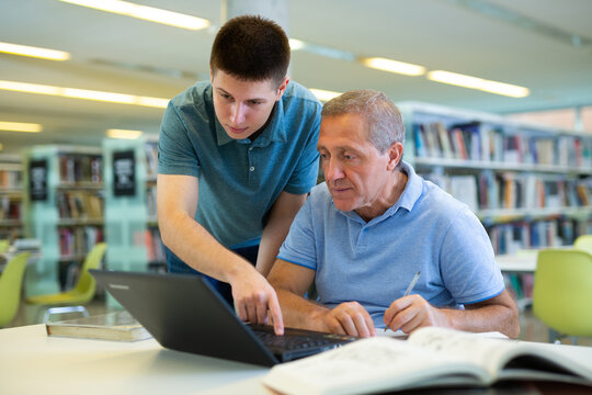 Young Guy Helping Older Man In Laptop Interface In Public Library