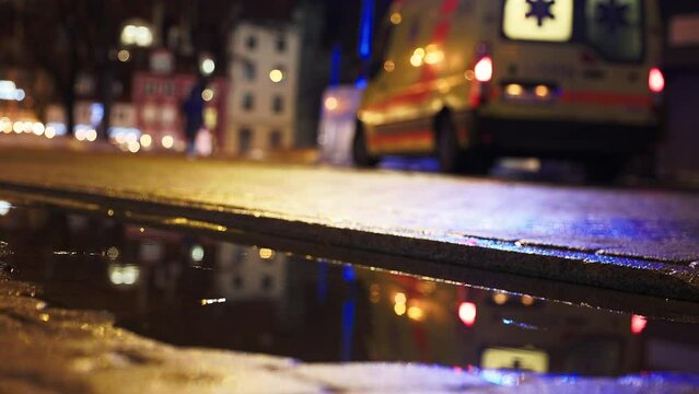 Revealing shot: An accident at night against a blurry ambulance with turned on flashing emergency siren lights in the rainy background