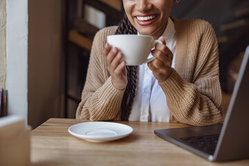 Focus on beautiful toothy smile of pretty ethnic woman holding cup with cappuccino during work on laptop in coffee shop