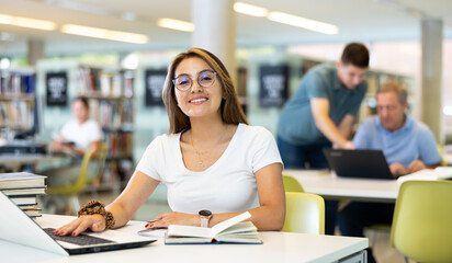 Confident student girl studying in the university library on a laptop, writes an important synopsis in a copybook