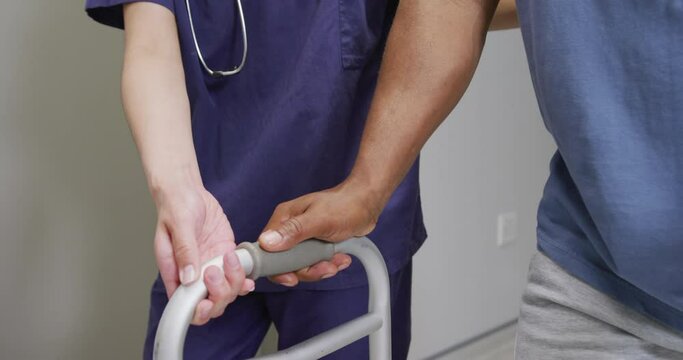 Midsection of diverse female doctor helping male patient use walking frame at hospital