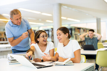 Mature man placing fingers on lips with shh near a table with laughing women using computer in the library