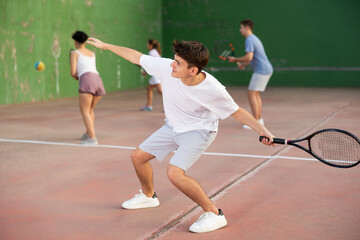 Concentrated young Latin American frontenis player swinging string racquet to hit ball on outdoor walled court on sunny summer day. Sport and active lifestyle concept