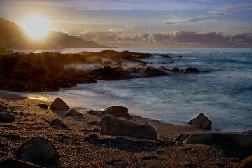 Long exposure landscape of the sunset on the beach. Seasape of waves breaking on the rocks of the Galician coast. Sunstar over the mountain. Baiona, Rias Baixas, Galicia, Spain