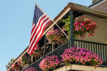 American flags hang off a second floor balcony of a historic building. The railings have baskets of pink and purple flowers hanging. The sky is blue and there are multiple windows in the old building.