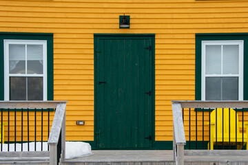 A vibrant yellow wooden exterior wall of a beach house with a solid green shutter door and two...