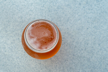 Top view of a glass of sour flavored beer with its head covered in froth. The liquid has an orange tint. There's a plastic cup cover on the beer on a composite plastic table top at a microbrewery. 