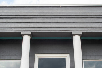 The roof section of an old grey colored cement business building wall. There are two white columns on either side of the door with two glass windows on the outside of the pillars. The sky is grey.
