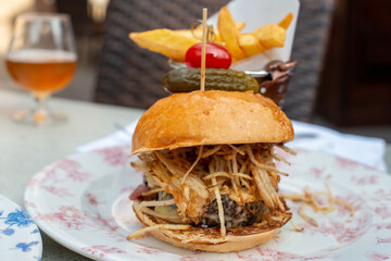 A hamburger with a pickle and tomato attached to the top bun on a blue and white plate with a copper cup filled with French fries. There's a pulled spicy pork sandwich on a plate on the table. 