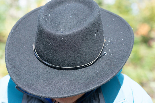A male wearing a wide brim black wool fedora style hat with a narrow leather sweatband. The man is wearing a pale blue shirt outdoors. The man's head is tilted downward. 