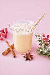 Glass of tasty eggnog cocktail, spices and Christmas decor on pink background