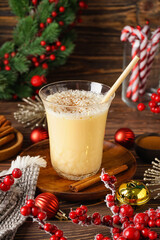 Glass of tasty eggnog cocktail, spices and beautiful Christmas decor on wooden background
