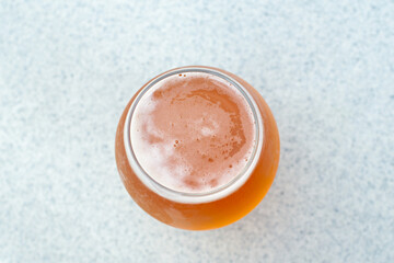 Top view of a glass of sour flavored beer with its head covered in froth. The liquid has an orange tint. There's a plastic cup cover on the beer on a composite plastic table top at a microbrewery. 