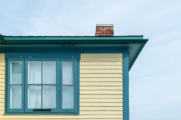 The exterior of a vintage yellow colored exterior wall with narrow wood cape cod clapboard siding with green trim. In the corner of the house is a wooden multi-pane window with lace white curtains.