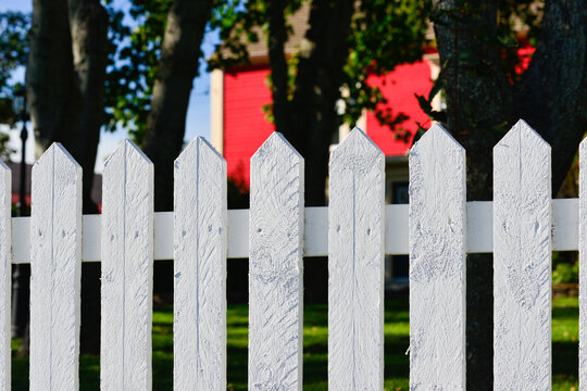 The exterior of a red wooden cape cod clapboard or batten board siding wall of a country style cottage with a white wooden picket fence in the foreground. There's a narrow horizontal line pattern.  