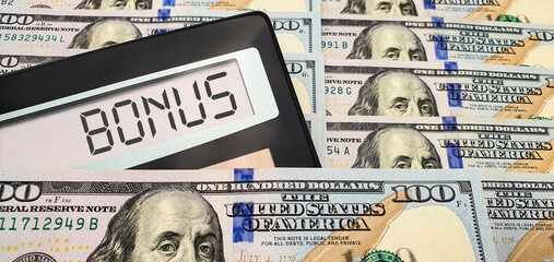 Calculator with the word "bonus" on the screen against the background of a large number of US 100 bills. The concept of the possibility of getting a big bonus