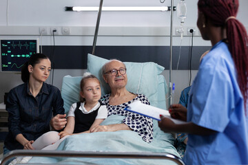Grandfather, close relatives receiving report of old man surgery. African american medical student attending bedridden elderly man discomfort. Medical staff noting senior symptoms.