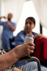 Close-up view of elderly woman in wheelchair holding bottle of pills for medical treatment. Nursing home nurse in scrubs assisting female patient, conversing and clarifying doubts.