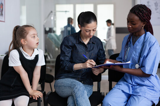 African American Doctor Filling Out Child Admission Paperwork To Pediatric Hospital. Young Woman Hospitalizing Sick Daughter In Medical Clinic. Nurse Attending Mother, Little Girl In Sanatorium Lobby.