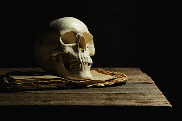 Human skull and old book on wooden table against black background, space for text