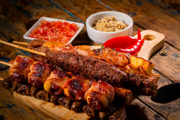 Espetinhos de churrasco em tábua de madeira com acompanhamentos, arroz, vinagrete, farofa e maionese