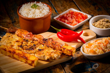 Espetinhos de churrasco em tábua de madeira com acompanhamentos, arroz, vinagrete, farofa e maionese © Vincius