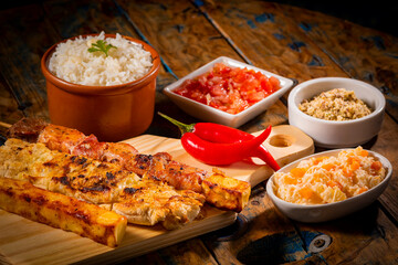 Fototapeta na wymiar Espetinhos de churrasco em tábua de madeira com acompanhamentos, arroz, vinagrete, farofa e maionese