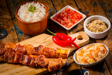 Espetinhos de churrasco em tábua de madeira com acompanhamentos, arroz, vinagrete, farofa e maionese