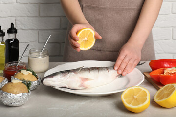 Woman squeezing lemon juice on fresh sea bass fish at light gray marble table, closeup