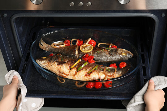 Woman taking out baking tray with sea bass fish and garnish from oven, closeup