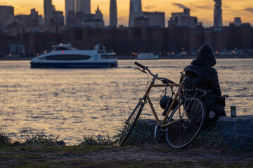 A New Yorker sitting by the river with her bicycle at sunset, the background is the skyline of Manhattan and ferry on east river. - obrazy, fototapety, plakaty