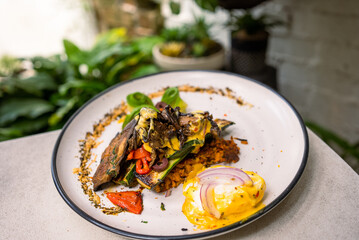 Stack of zucchini, eggplant, and capsicum pan-fried with potato fritters, poached egg, red onion, and cheese sauce on the side. Decorated with chia seeds and avocado and served with a cappuccino.