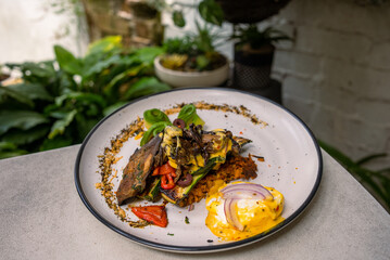 Stack of zucchini, eggplant, and capsicum pan-fried with potato fritters, poached egg, red onion, and cheese sauce on the side. Decorated with chia seeds and avocado and served with a cappuccino.