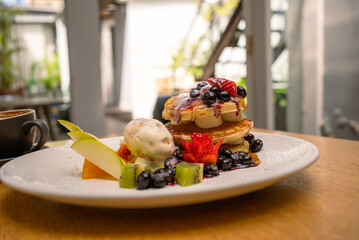 A plate of fluffy pancakes with delicious berries, a slice of orange, apples, maple syrup, and ice cream for breakfast on a light garden background with a cappuccino on the side