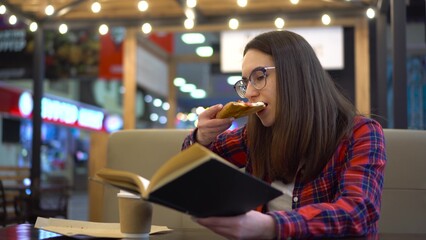 A young woman is reading a book in a cozy cafe. A woman sits at a table and eats pizza in the food court. Girl in a red shirt with glasses.